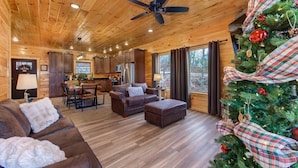 A log cabin interior with a dining area, a living room with a sofa and ottoman, a kitchen, and a decorated Christmas tree. - Pine Haven Cabin (Sevierville)