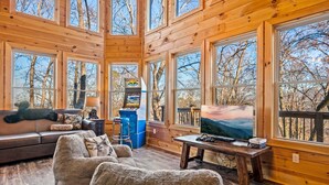 A wooden cabin interior with a sofa, armchair, TV, and arcade machine. - Z - Honey Bear Treehouse (Gatlinburg)