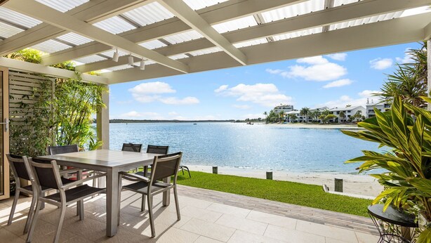 Eine Terrasse im Freien mit Tisch und Stühlen, Blick auf das Wasser und ein Gebäude im Hintergrund.