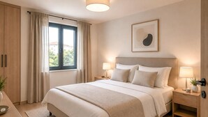 Ein Schlafzimmer mit einem Bett, zwei Nachttischen mit Lampen, einem Fenster mit Vorhängen und einem an der Wand hängenden Kunstwerk.
