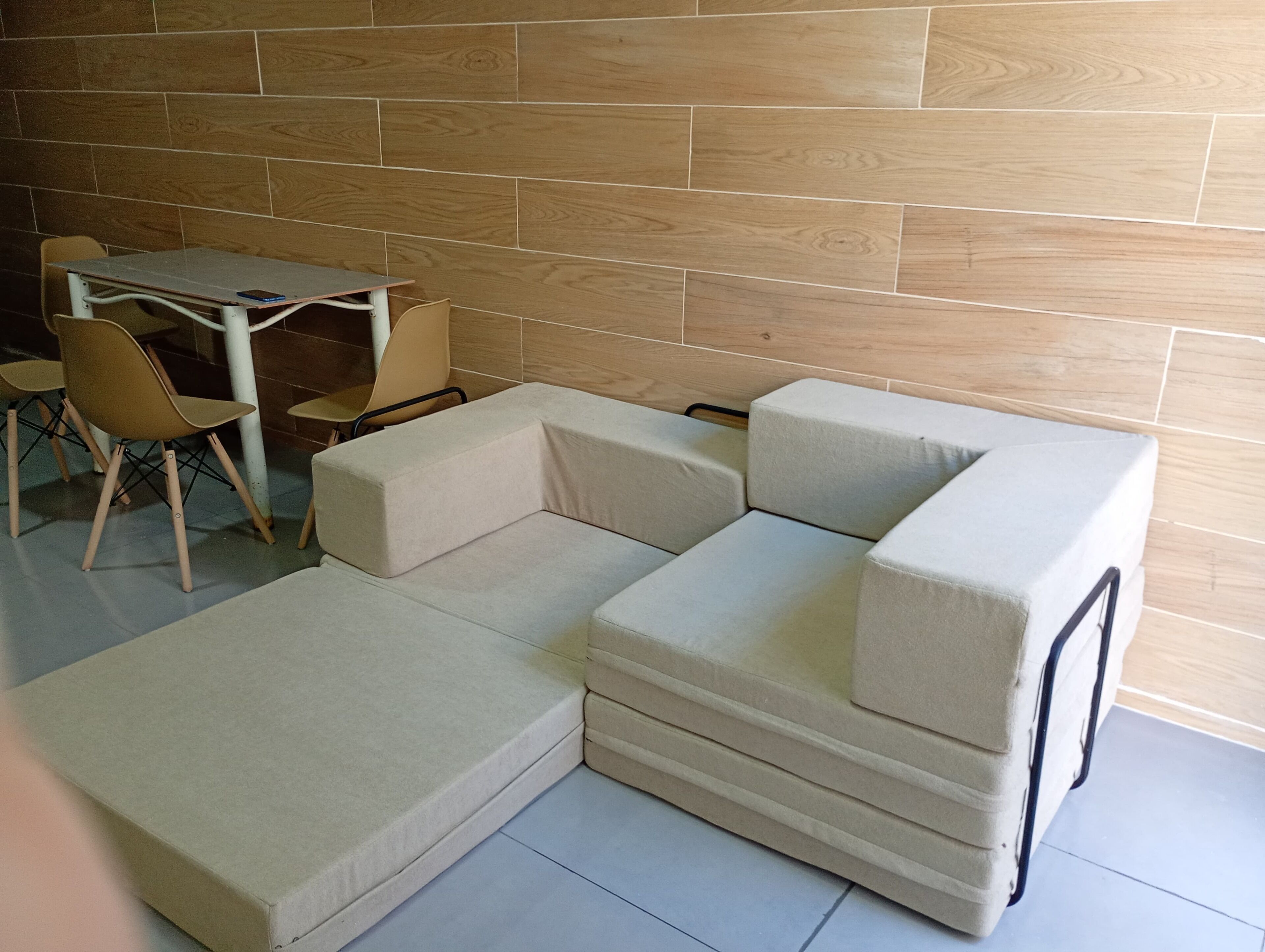 Un canapé modulable, une petite table et deux chaises dans une pièce dont le sol est en bois et un des murs est recouvert de carreaux.