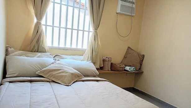 Un dormitorio con cama, cortinas, un equipo de aire acondicionado y un estante con objetos.