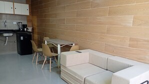 Una kitchenette moderna con cafetera, refrigerador pequeño y lavamanos. Hay una zona de comedor con mesa y sillas, y un sofá con cojín.