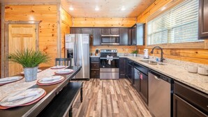 A modern kitchen with wooden walls, stainless steel appliances, and a dining area with a table set for four. - Magical Meadow (Sevierville)
