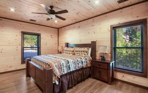 A bedroom with a wooden ceiling, a bed with patterned bedding, a nightstand, a lamp, and a window with blinds. - Misty Shadows (Sevierville)