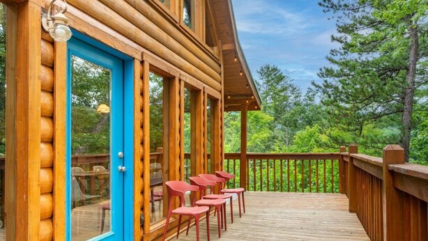 Um deck de madeira com uma porta azul, cadeiras vermelhas e vista para árvores.