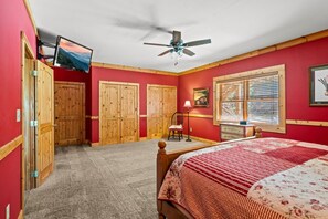 Quarto com cama, ventoinha no teto, TV fixada na parede, portas de madeira, janela com persianas e luminária.