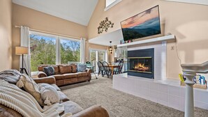 A living room with a fireplace, a sofa, a dining area, and a large window with a view of trees. - Smoky Dreams 2 (Gatlinburg)