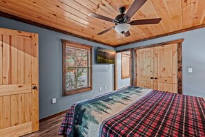 A bedroom with a bed, wooden ceiling, a ceiling fan, a window with a view, and a door. - Mountain Vibes (Gatlinburg)