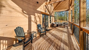 Eine Holzterrasse mit Adirondack-Stühlen und einem Tisch, ein überdachter Bereich mit Geländer und Blick auf Bäume.