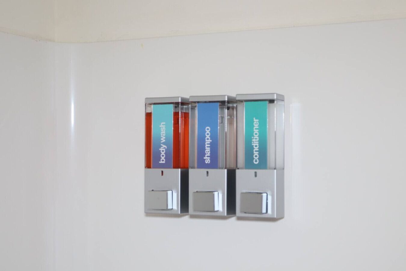 Dispensador montado en la pared con tres compartimentos etiquetados como gel de ducha, champú y acondicionador.
