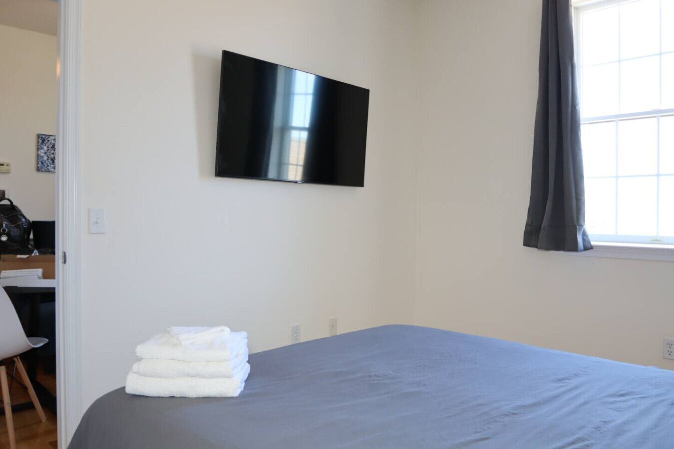 A bedroom with a bed, a TV mounted on the wall, a window with curtains, and a desk with a chair.