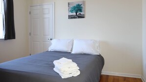 Una habitació amb un llit, almoines blanques, una tovallola blanca doblegada, una porta i un quadre penjat a la paret.