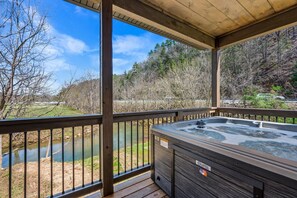 A hot tub on a wooden deck with a view of a river and forest. - Cabin By The Creek (Sevierville)