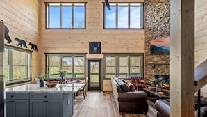 A spacious interior with a kitchen, dining area, and living room, featuring wooden walls, large windows, and a stone fireplace. - Cabin By The Creek (Sevierville)