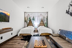Ein Zimmer mit zwei Betten, einem Schlafsofa, einem Schachbrett und einem Waldgemälde.