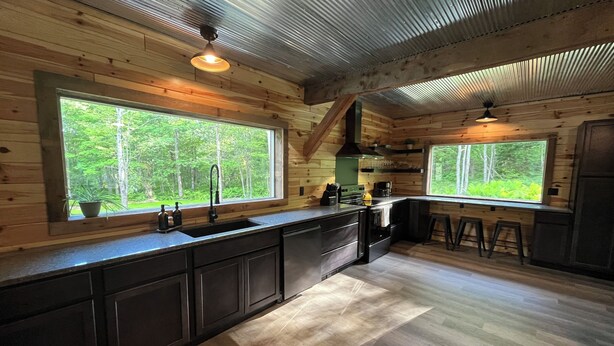 A wooden cabin interior with a kitchen, large windows, and a view of the forest.