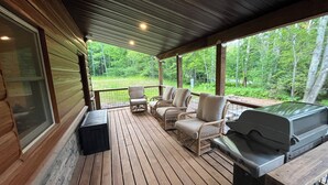 A covered deck with wooden flooring, wicker chairs, a grill, and a view of a forested area.