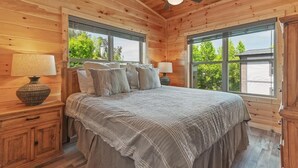 Ein Schlafzimmer in einer Holzhütte mit einem Bett, zwei Fenstern, einem Deckenventilator und einem Nachttisch mit Lampe.