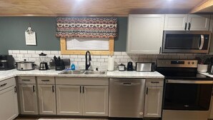 A modern kitchen with stainless steel appliances, a built-in dishwasher, and a window with patterned curtains. - Cozy Hollow Cabin (Sevierville)