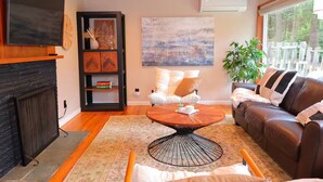 Ein Wohnzimmer mit Kamin, Fernseher, Bücherregal, einem Gemälde, einer Couch, einem Couchtisch und einer Pflanze.