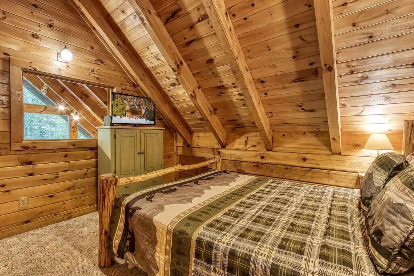 Un dormitorio en el ático acogedor, con techo inclinado de madera, una cama con colcha escocesa, un armario verde y un televisor en una estantería.