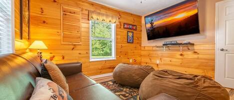 A cozy living room with a leather couch, a flat-screen TV, wooden walls, a ceiling fan, and a window with a view of greenery.