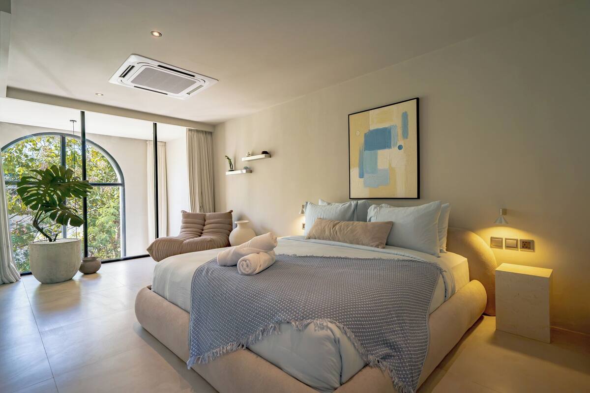 A modern bedroom with a large bed, a potted plant, and a view through a large window.