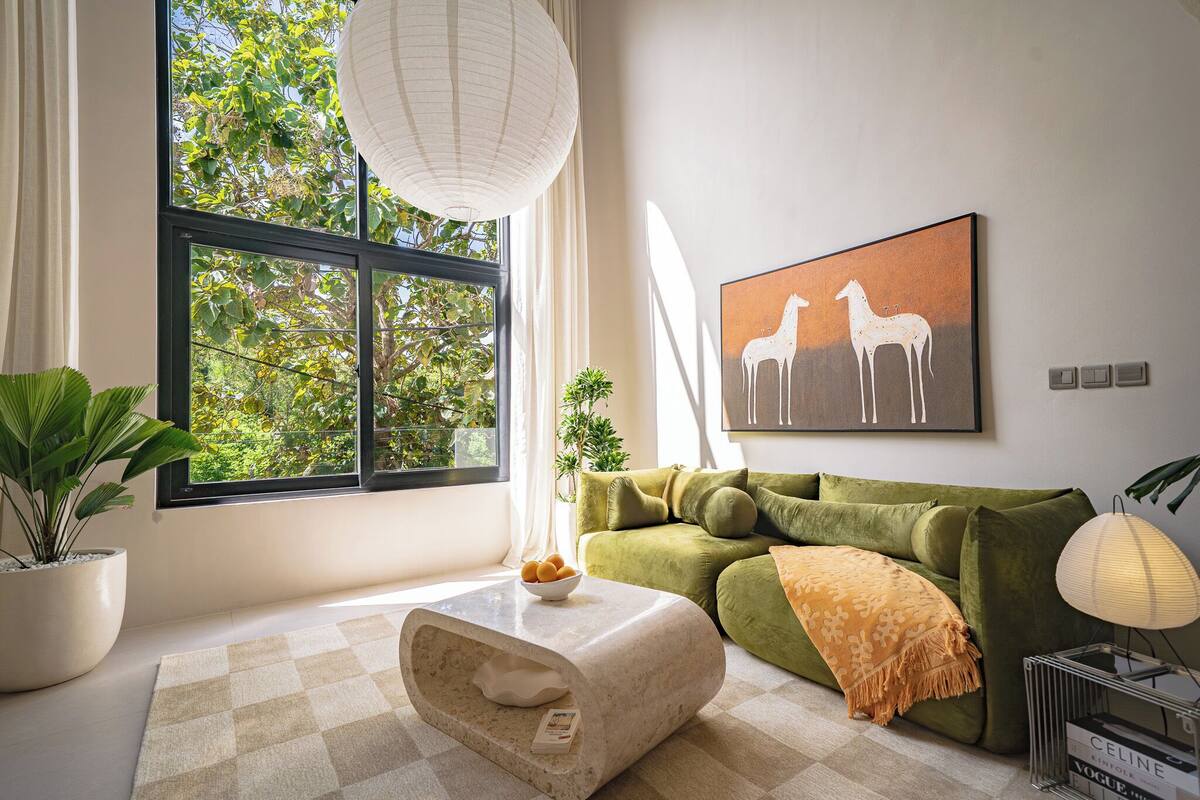 A modern living room with a green sofa, a white coffee table, a large window with a view of trees, and a painting of horses on the wall.