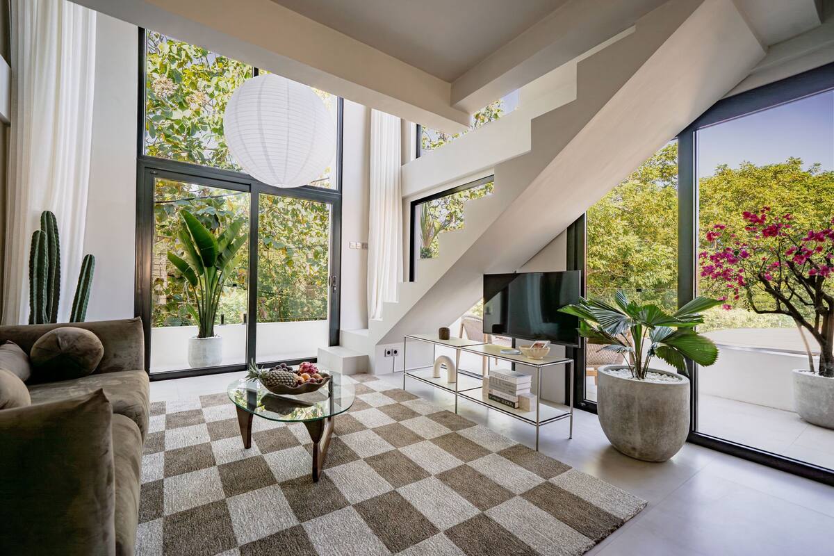 A modern living room with a checkered rug, a glass coffee table, a sofa, a TV, and large windows with plants.