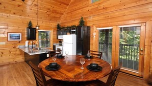 Interior d'una cabana de fusta amb una taula de dinar per a quatre, una cuina amb nevera i fogons, i una porta corredissa de vidre a l'exterior.
