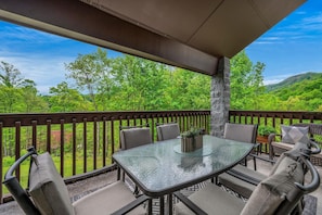 A covered outdoor patio with a glass table and chairs, a stone pillar, and a view of green trees and hills. - Gracious Oasis (Gatlinburg)
