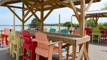 A wooden deck with a long table and colorful chairs, overlooking a body of water.