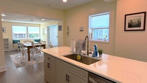 Una cocina moderna con lavavajillas de acero inoxidable, un lavamanos y un grifo, y una ventana encima. Se ve una zona de comedor con una mesa y sillas al fondo.