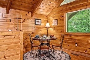 A wooden cabin interior with a round dining table, metal chairs, a lamp, framed pictures, and a window with a view of trees. - Mountain Dream (Sevierville)