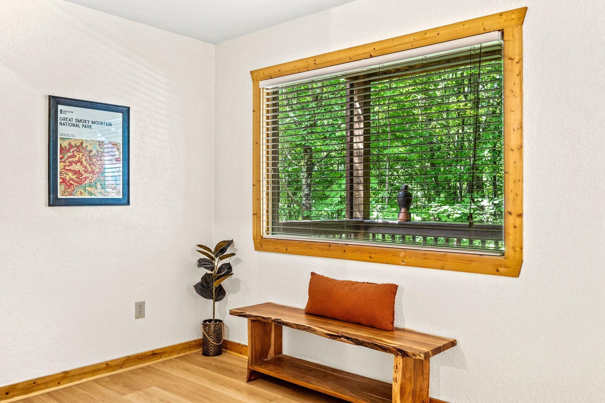 A room with a wooden bench, a window with blinds, a framed map on the wall, and a potted plant.
