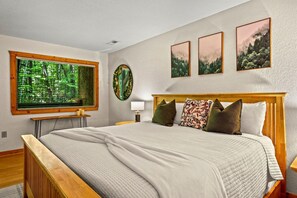A bedroom with a large bed, a window with a view of greenery, a round mirror, and three framed pictures on the wall. - Mountain Dream (Sevierville)