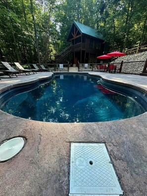 Ein Schwimmbecken mit Holzterrasse, Liegestühlen und einer Hütte in einem bewaldeten Gebiet.