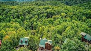 A forested area with several buildings, including cabins with green roofs, surrounded by dense trees and distant mountains. - Away in the Mountains (Sevierville)