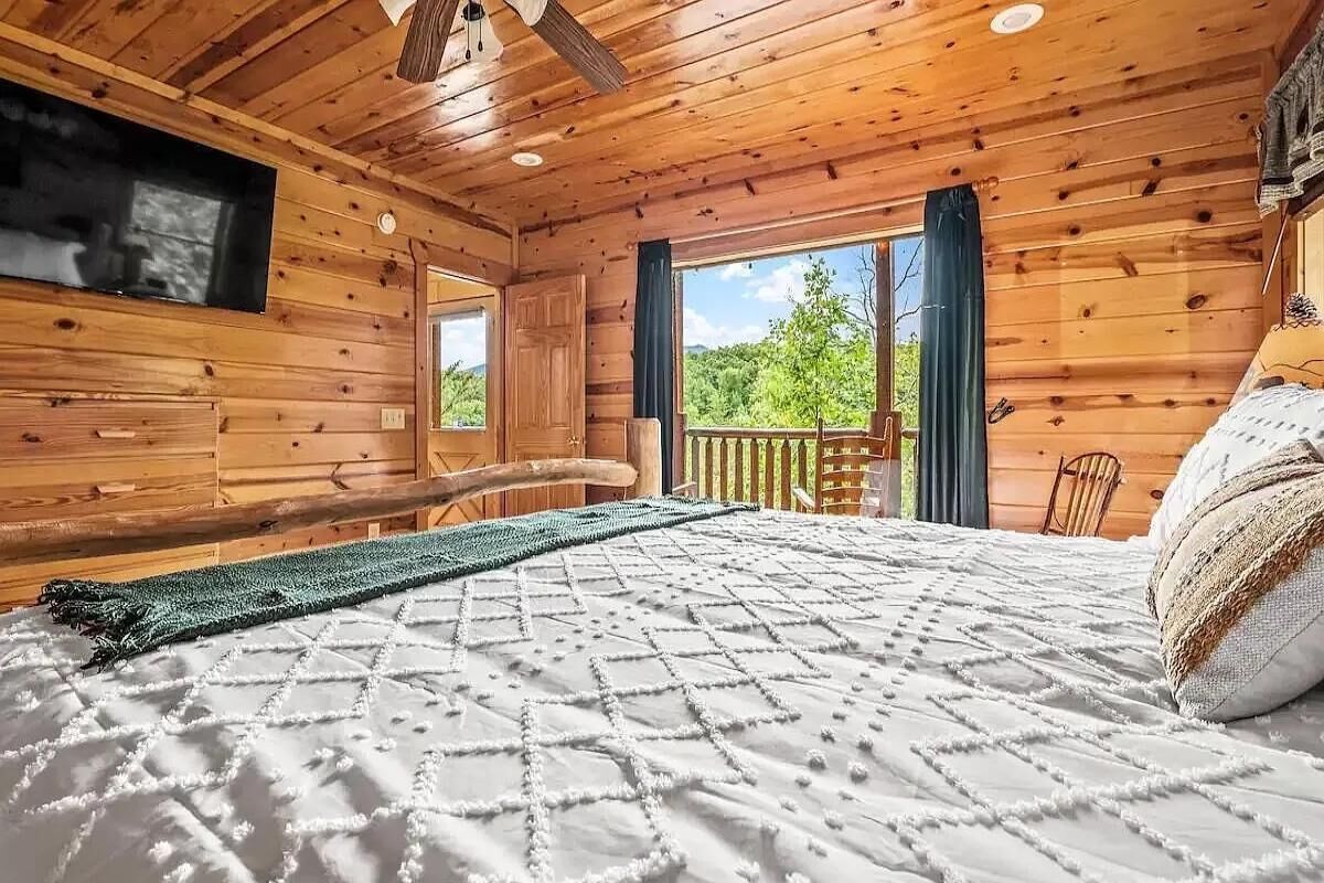 Een slaapkamer in een houten hut met een bed, een televisie, een balkon en uitzicht op de omgeving.