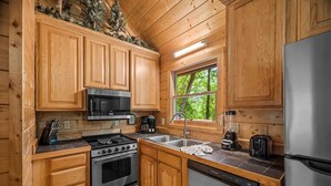 Eine Holzhütte mit Küche, inklusive Herd, Mikrowelle, Spüle, Kühlschrank und einem Fenster mit Blick auf Bäume.