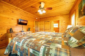 A wooden cabin bedroom with a bed, ceiling fan, wooden walls, a TV, and a window. - Nicoha (Sevierville)