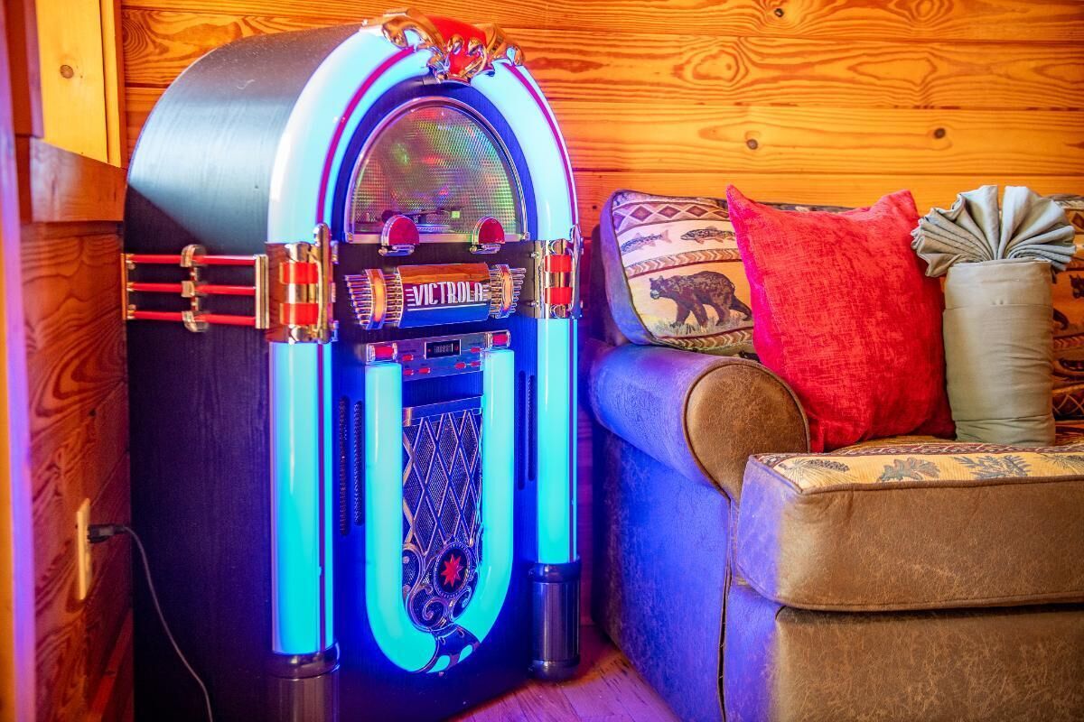 Ein Jukebox mit runder Oberseite und einer verzierten Frontblende, ein braunes Sofa mit gemusterten Kissen und eine Holzwand im Hintergrund.