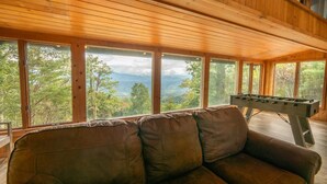 Ein Holzinnenraum mit einer Couch und einem Kicker, große Fenster mit Blick auf Bäume und Berge.
