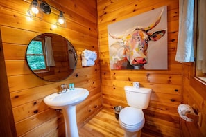 Ein Badezimmer in einer Holzhütte mit einem runden Spiegel, einem weißen Waschbecken, einer Toilette und einem Bild einer Kuh an der Wand.