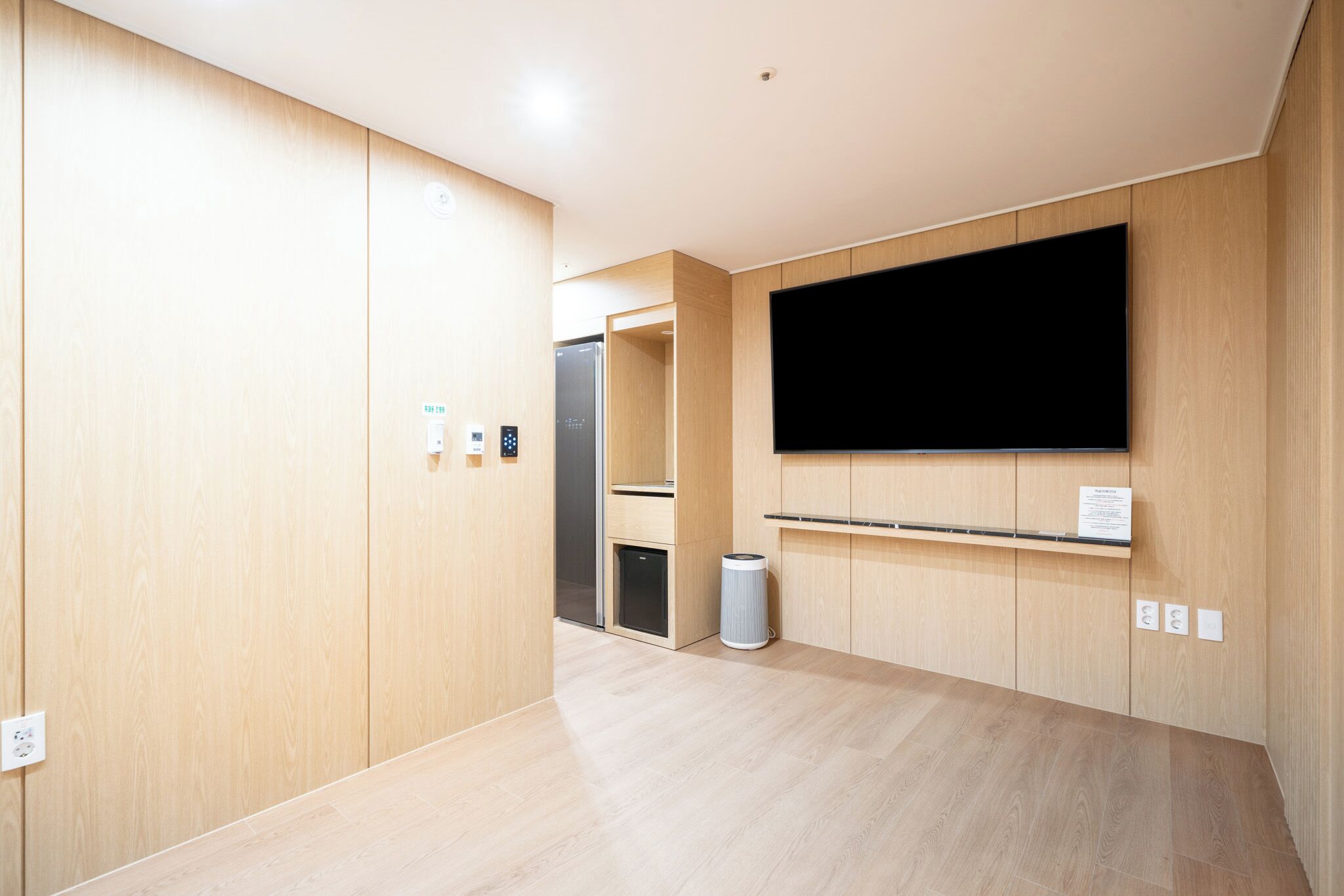 A modern hotel room with wooden panel walls, a flat-screen TV mounted on the wall, a small trash bin, and a door leading to a bathroom.