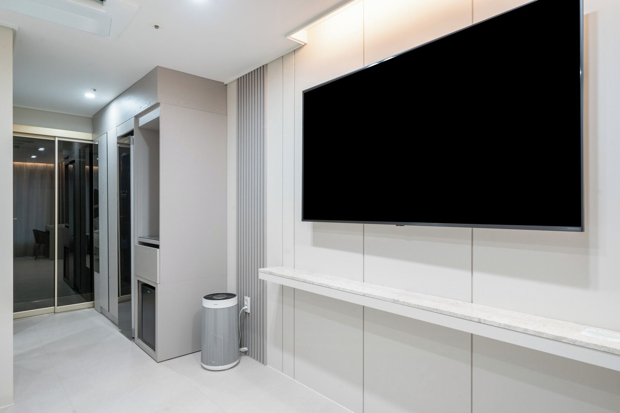 A modern hotel room with a large flat-screen TV mounted on the wall, a glass partition, and a small trash bin.