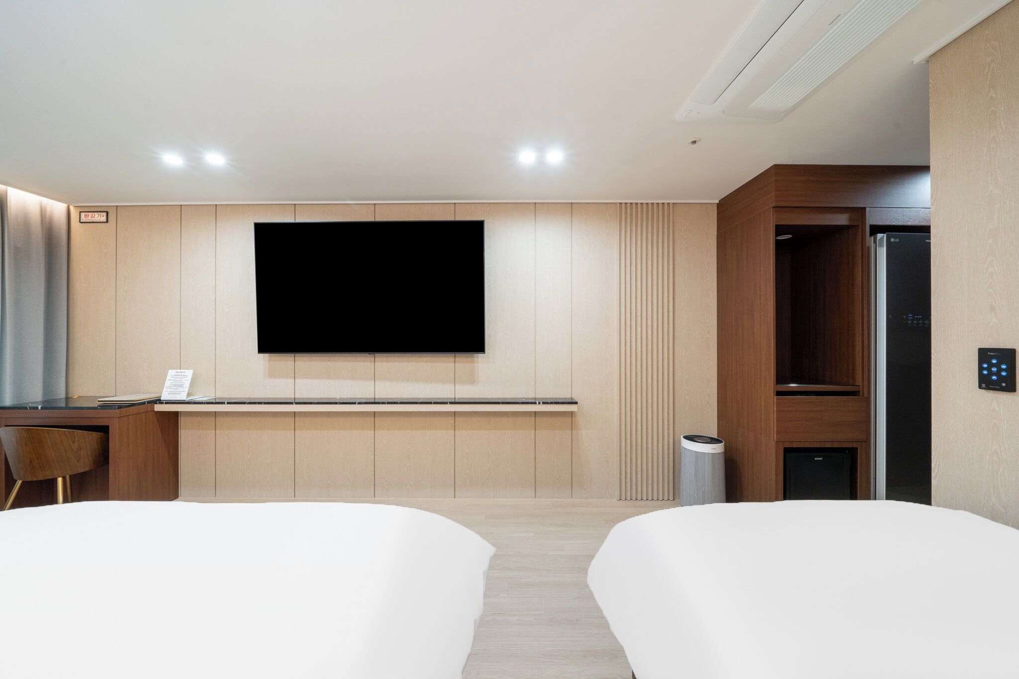 Una habitación de hotel moderna con televisión de pantalla plana en la pared, un escritorio con una silla, dos camas, un cubo de basura y un refrigerador empotrado.