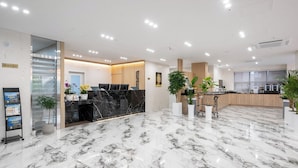 Un vestíbulo de hotel con piso de mármol, mostrador de recepción, plantas en macetas y un quiosco de información digital.