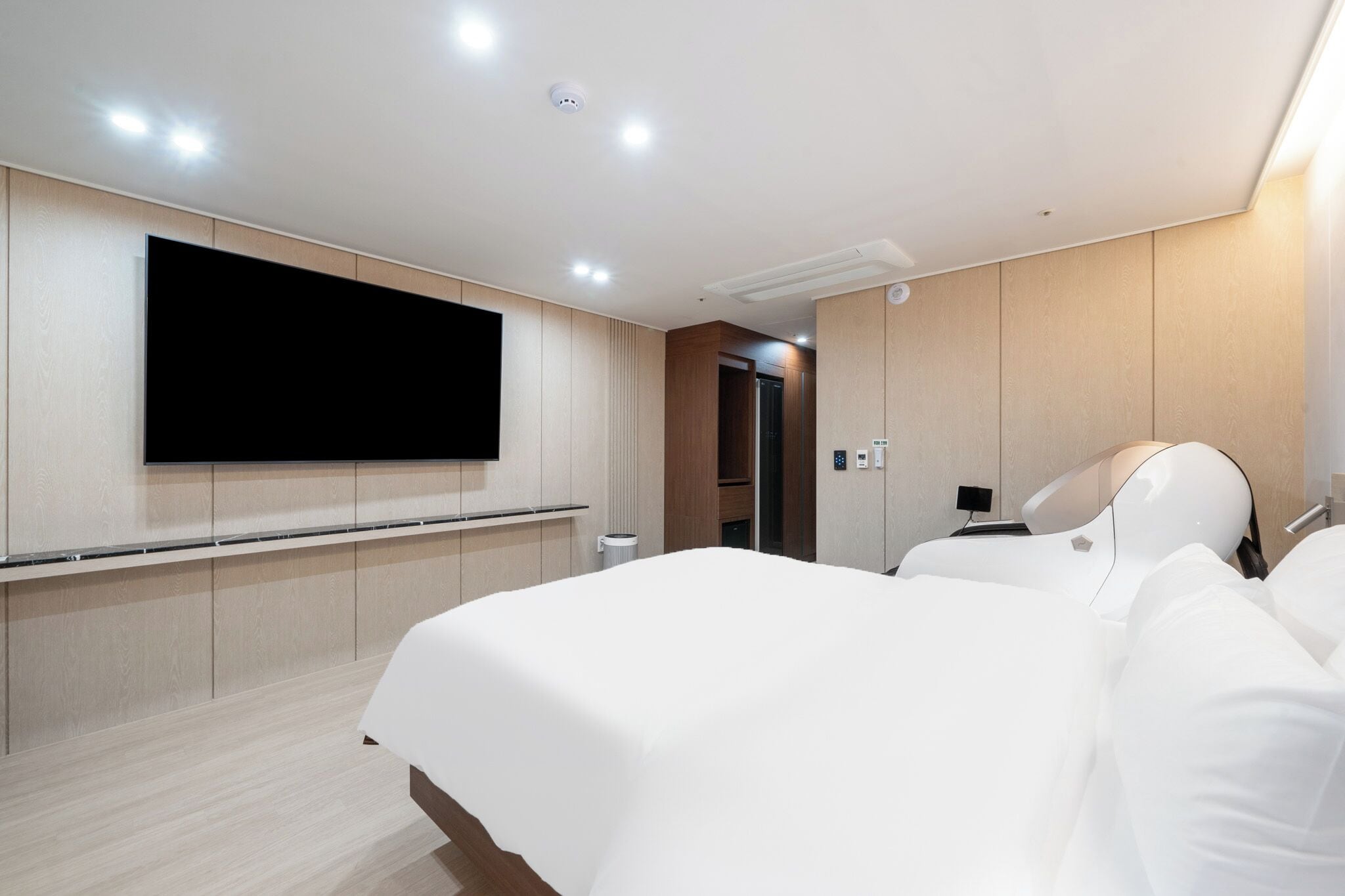 Une chambre d’hôtel moderne dotée d’un grand téléviseur à écran plat fixé au mur, d’un lit avec du linge de lit blanc et d’une table de chevet.